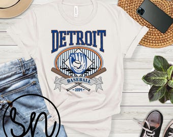 May include: Off-white t-shirt with a Detroit Baseball graphic. The design features the word "DETROIT" in blue, an oval with a baseball glove and ball, crossed bats, and the text "BASEBALL 1894".