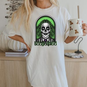 May include: Cream-colored t-shirt featuring a graphic of a skull with green hair and the word "Beetlejuice" in green. The skull is set against a green background within a black dotted frame. The shirt is a casual style.