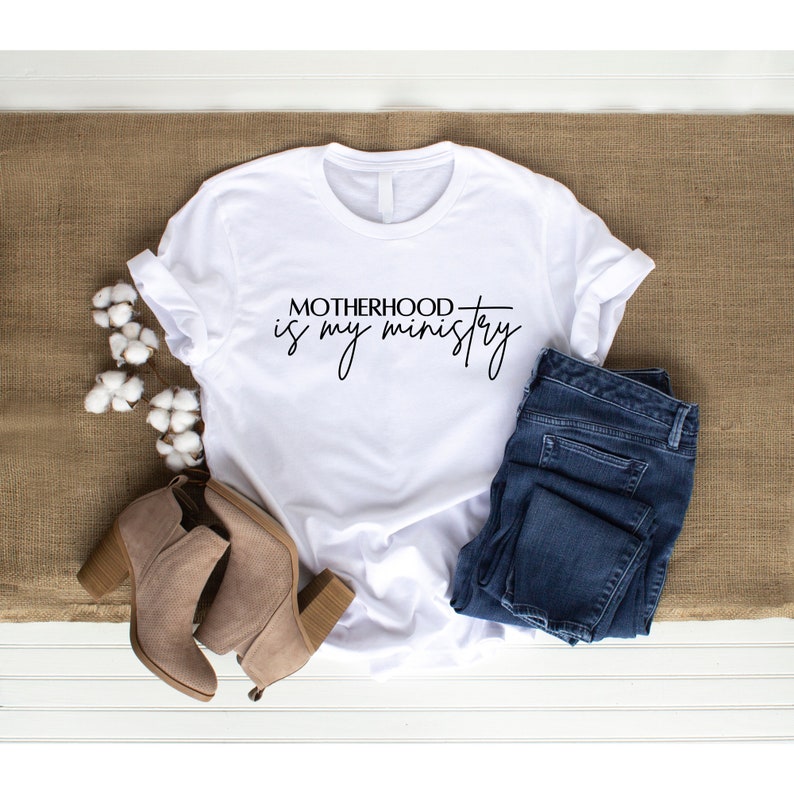 May include: White t-shirt with the text "Motherhood is my ministry" in black script font. The shirt is laid flat on a brown burlap surface with a pair of brown suede ankle boots and blue jeans.