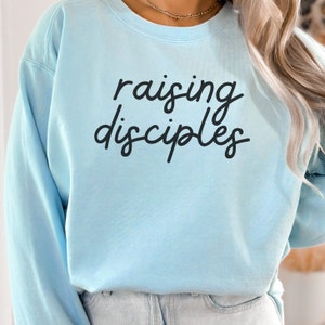 Könnte beinhalten: Hellblauer Pullover mit dem Text "raising disciples" in schwarzer Schrift.