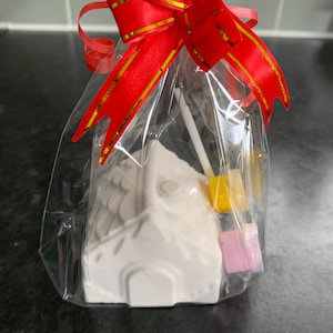 May include: A white ceramic house, packaged in clear cellophane and tied with a red ribbon with gold detailing. The package contains small yellow and pink items, possibly sweets or decorations.