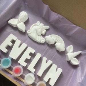 May include: A paint-your-own craft kit featuring the name "EVELYN" in white ceramic letters, along with a unicorn, mermaid, rainbow, and unicorn horn. The kit includes six small paint pots with various colours.