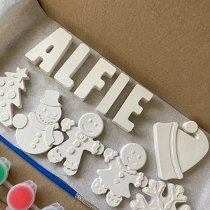Puede incluir: Un kit de manualidades con el nombre "ALFIE" en letras de bloque, junto con formas navideñas sin pintar: un árbol de Navidad, un muñeco de nieve, hombrecitos de jengibre, un gorro y un copo de nieve. Incluye un pincel y botes de pintura.