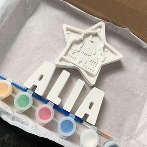 May include: A white ceramic star with a mosque design and the name "ALIA" spelled out in white ceramic letters. The set comes with six small paint pots in various colours and a paintbrush.
