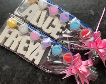 Könnte beinhalten: Zwei Sätze weißer Holzbuchstaben, die die Namen "Alice" und "Freya" buchstabieren, werden mit Farbtöpfen und Pinseln verpackt. Die Sets sind in Klarsichtfolie verpackt und mit einem rosa Band mit goldenen Akzenten gebunden.
