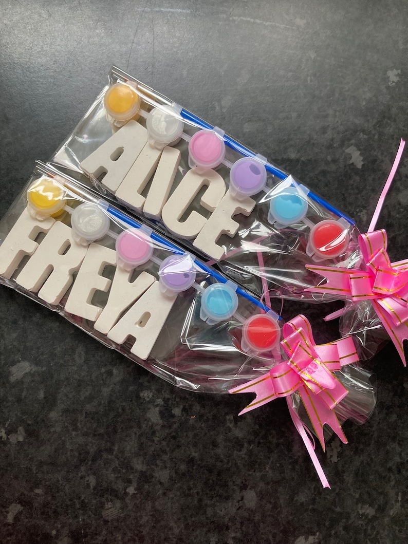 May include: Two sets of white wooden letters spelling out the names "Alice" and "Freya" are packaged with paint pots and brushes. The sets are wrapped in clear plastic and tied with a pink ribbon with gold accents.