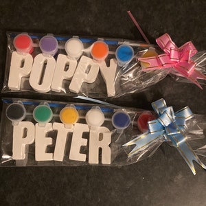 May include: Two sets of paint-your-own wooden letters, one set spelling "POPPY" and the other spelling "PETER". Each set comes with six small paint pots in different colors, a paintbrush, and a clear plastic bag with a bow.