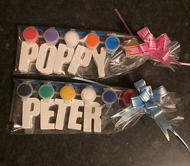May include: Two sets of paint-your-own wooden letters, one set spelling "POPPY" and the other spelling "PETER". Each set comes with six small paint pots in different colors, a paintbrush, and a clear plastic bag with a bow.