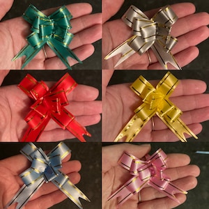 May include: Six different colors of gift bows with gold trim. The bows are made of satin ribbon and have a loop in the center. The bows are arranged in a grid pattern.