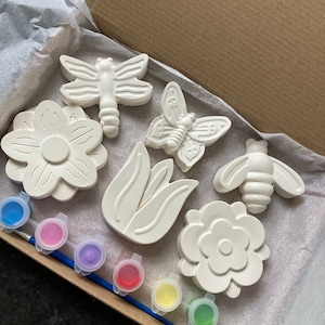 May include: A set of six white ceramic flower and insect shapes for painting. The set includes a dragonfly, butterfly, bee, tulip, and two flowers. The set also includes six small paint pots in blue, pink, purple, red, yellow, and green.