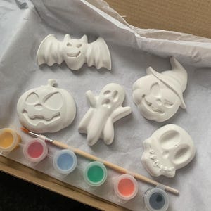 May include: A Halloween-themed craft kit featuring unpainted ceramic figures of a bat, pumpkin, ghost, witch, and skull. Includes a paintbrush and six paint pots with various colors, all inside a cardboard box.