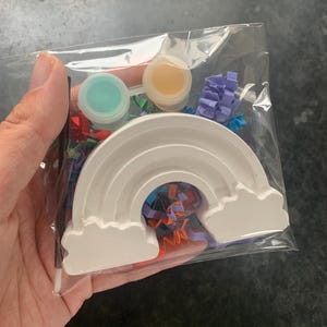 May include: A clear plastic bag contains a white rainbow-shaped craft item with two attached cloud shapes. The bag also holds two small paint pots, one teal and one yellow, and colorful confetti.