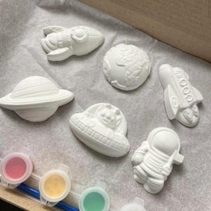 May include: Set of six white ceramic space-themed ornaments, including a rocket, astronaut, UFO, planet, and two stars. The ornaments are ready to be painted.