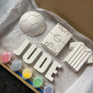May include: A white paint-your-own craft kit with the name "JUDE" spelled out in large letters. The kit includes a football, a football shirt, a football pitch, and six small paint pots.