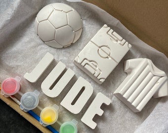 Könnte beinhalten: Ein weißes Bemal-Set zum Selbermachen mit dem Namen "JUDE" in großen Buchstaben. Das Set enthält einen Fußball, ein Fußballtrikot, einen Fußballplatz und sechs kleine Farbtöpfe.