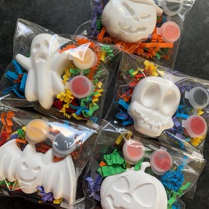 May include: Five Halloween-themed paint-your-own kits. Each kit includes a white ceramic figure of a ghost, witch, skull, bat, or jack-o'-lantern, paint pots, and colorful paper filler, all packaged in a clear plastic bag.