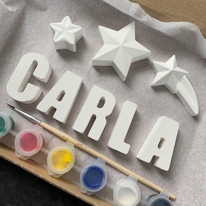 Peut inclure: Un kit de bricolage DIY comprenant le nom "CARLA" en blanc, ainsi que trois étoiles blanches et une étoile filante. Le kit comprend six petits pots de peinture en vert, rouge, jaune, bleu, blanc et noir.