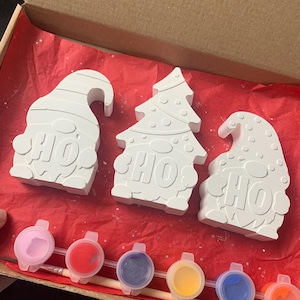 May include: A craft kit featuring three white plaster Christmas ornaments: two gnomes and a Christmas tree, each with the word "HO" on them. The kit includes a paintbrush and several small paint pots with various colors, all inside a cardboard box.