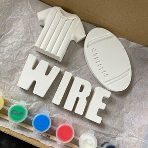 May include: A craft kit featuring a white ceramic shirt, a rugby ball shape, and the word "WIRE". The kit includes paint pots in various colours and paintbrushes, all inside a cardboard box. The items are unpainted and ready for customisation.