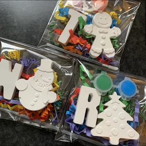 May include: Three clear bags containing craft kits. Each bag has a white ceramic shape, colourful paper shreds, and paint pots. Shapes include a snowman, a gingerbread man, a Christmas tree, and letters.