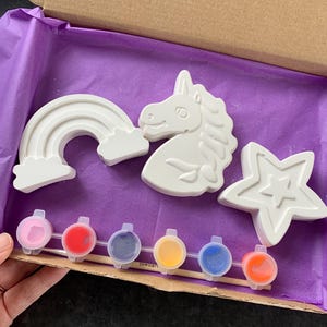 May include: A craft kit featuring plaster figures of a rainbow, unicorn head, and star. The kit includes six small paint pots in various colours and a wooden stick. The items are arranged inside a cardboard box with purple tissue paper.