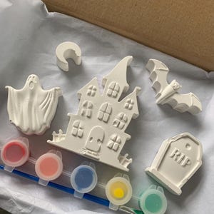 May include: A Halloween-themed paint kit featuring a haunted house, ghost, bat, tombstone, and crescent moon. Includes six paint pots with various colors and a paintbrush. The kit is presented in a cardboard box with white tissue paper.