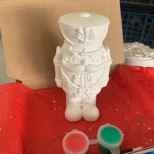 May include: A white, unpainted nutcracker figurine with detailed uniform features, ready for painting. Includes small pots of red and green paint, and a paintbrush. The figurine is on a red surface, with a cardboard backing.