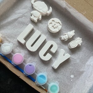 May include: A craft kit with white paint-your-own ceramic pieces, including the name "Lucy", a pineapple, a flower, and a cartoon character. The kit also includes five small pots of paint in various colors.