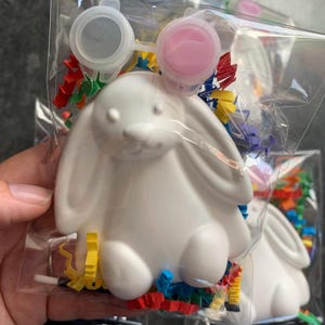 May include: A white bunny-shaped paint-your-own craft kit, packaged in a clear plastic bag. The kit includes paint pots with grey and pink paint, colourful confetti, and a small paintbrush. The bunny is approximately 13 cm tall.
