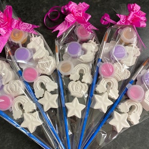 Könnte beinhalten: Vier durchsichtige Plastiktüten gefüllt mit Bastelbedarf zum Malen. Jede Tüte enthält einen kleinen Pinsel, ein Einhorn, eine Krone, einen Stern, einen Regenbogen und vier kleine Töpfe mit Farbe. Die Tüten sind mit rosa Schleifen gebunden.