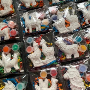 May include: A set of 10 paint your own llama kits. Each kit includes a white ceramic llama figurine, paint pots, and a paintbrush. The llamas are in various poses, including standing, sitting, and with a cactus. The kits are packaged in clear plastic bags with colorful tissue paper.