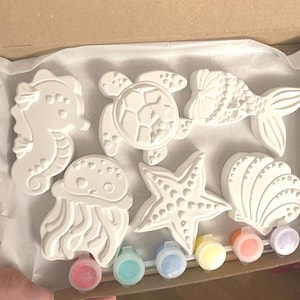 Könnte beinhalten: Ein Bastelset mit sechs weißen Meereskreaturen aus Gips: ein Seepferdchen, eine Schildkröte, ein Fisch, eine Qualle, ein Seestern und eine Muschel. Enthält sechs kleine Farbtöpfe in verschiedenen Farben. Das Set ist in einem Karton verpackt.