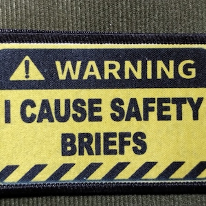 May include: A rectangular patch with a black border and the words "WARNING I CAUSE SAFETY BRIEFS" in black text on a yellow background. A black and yellow striped design is at the bottom, and a yellow exclamation point is at the top.