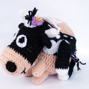 May include: A crocheted plush toy cow with black and white spots, wearing a pink flower on its head. The cow is riding on the back of a larger crocheted plush toy with a light brown body and black eyes.