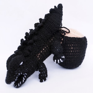 Può includere: Giocattolo di dinosauro in crochet nero con denti bianchi. Il dinosauro è in posizione accovacciata con la testa abbassata.