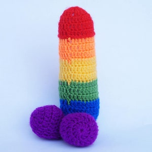 May include: A handmade crochet sculpture in the shape of a penis, featuring a rainbow color scheme. The head is red, transitioning through orange, yellow, green, and blue. Two purple crochet spheres are positioned below.
