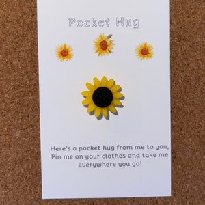 Puede incluir: Un girasol amarillo con un centro negro, con el texto "Pocket Hug" encima. El texto debajo del girasol dice "Here's a pocket hug from me to you, Pin me on your clothes and take me everywhere you go!"