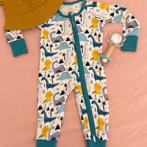 May include: A white baby onesie with a teal zipper and teal trim. The onesie has a pattern of blue, yellow, and gray dinosaurs. The onesie is laying on a pink surface.