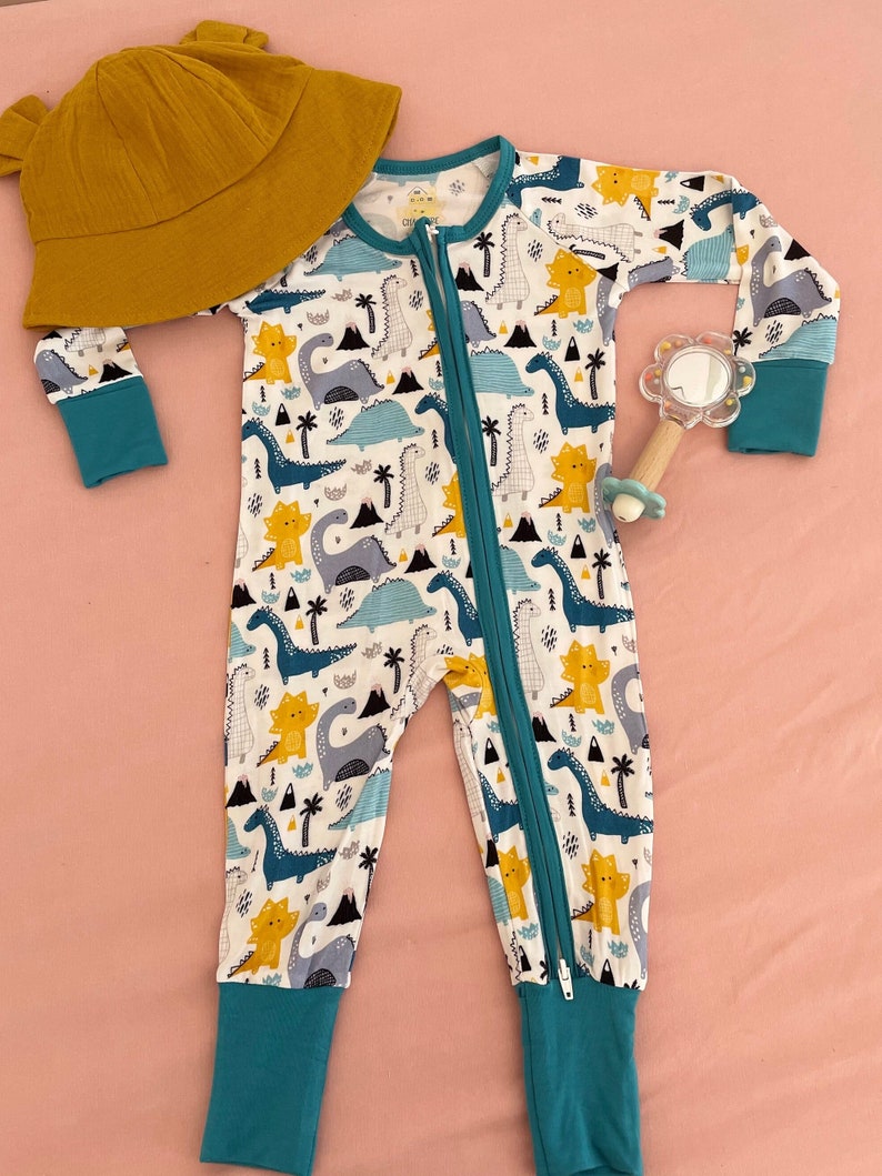 May include: A white baby onesie with a teal zipper and teal trim. The onesie has a pattern of blue, yellow, and gray dinosaurs. The onesie is laying on a pink surface.