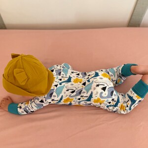 May include: A baby wearing a yellow hat with ears and a white onesie with a blue, yellow, and green dinosaur print.