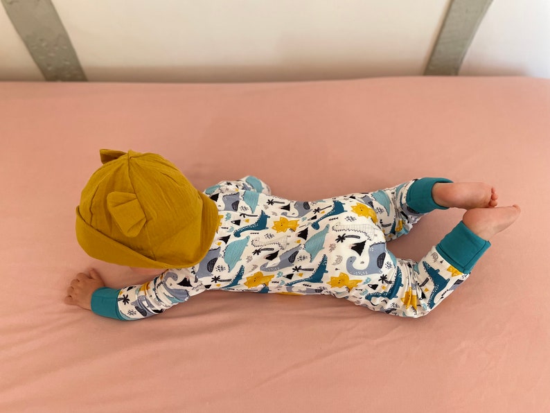 May include: A baby wearing a yellow hat with ears and a white onesie with a blue, yellow, and green dinosaur print.