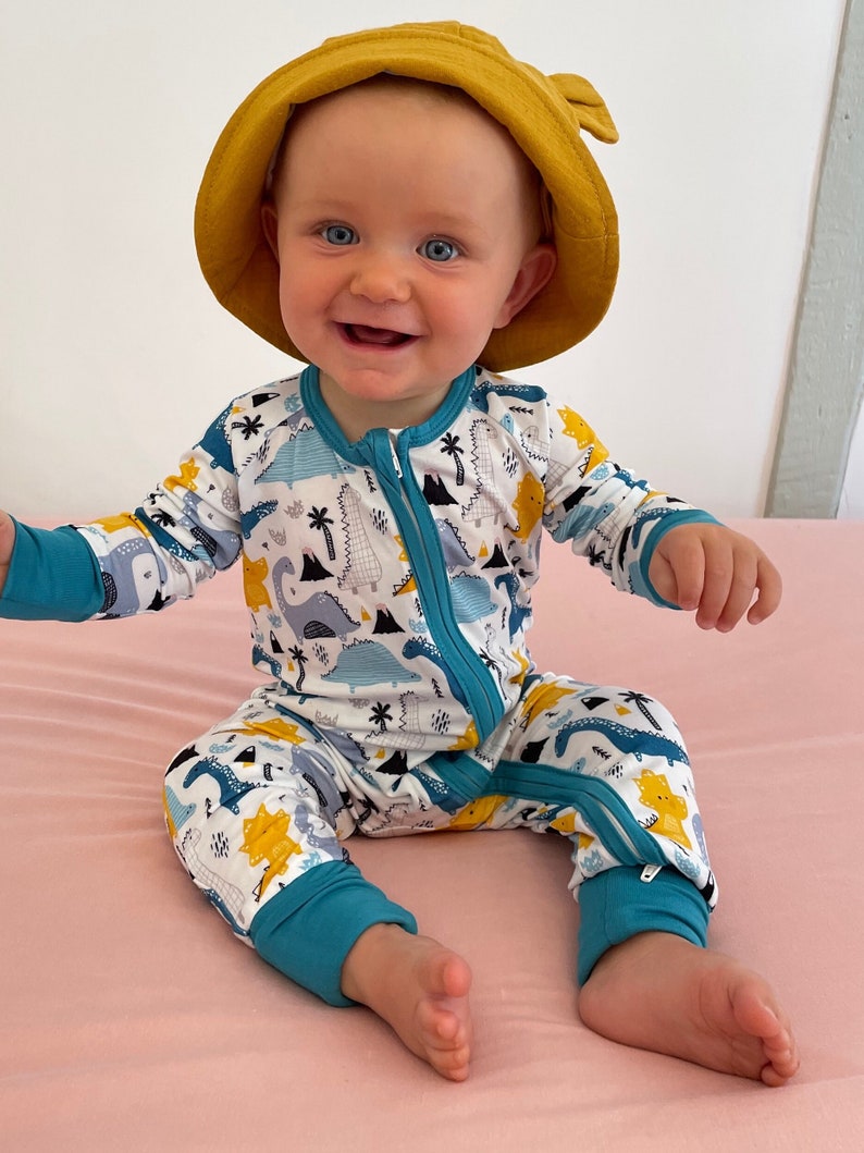 May include: A baby wearing a white and blue dinosaur print zippered jumpsuit and a yellow sun hat. The jumpsuit has a turquoise zipper and turquoise trim around the cuffs and legs.