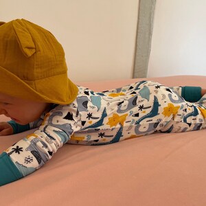 May include: A baby wearing a yellow sun hat and a white long-sleeved onesie with a dinosaur print. The onesie has blue, green, and yellow dinosaurs on it.