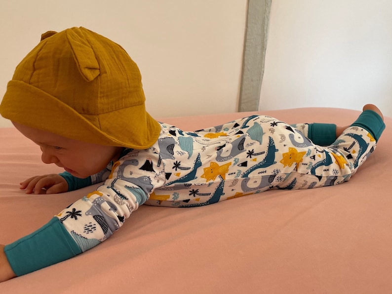 May include: A baby wearing a yellow sun hat and a white long-sleeved onesie with a dinosaur print. The onesie has blue, green, and yellow dinosaurs on it.
