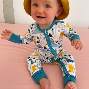 May include: A baby wearing a blue and white dinosaur print onesie with a yellow hooded hat. The onesie has a zipper down the front and blue cuffs on the sleeves and legs.