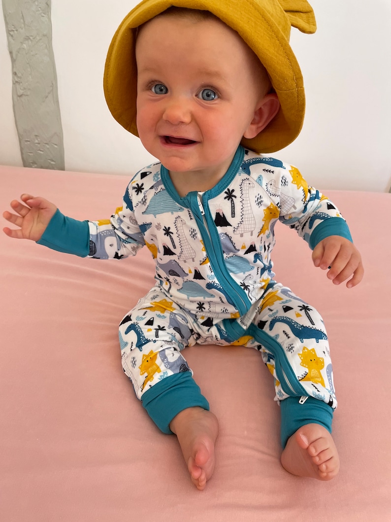 May include: A baby wearing a blue and white dinosaur print onesie with a yellow hooded hat. The onesie has a zipper down the front and blue cuffs on the sleeves and legs.