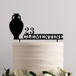 Op de afbeelding: Een zwarte taarttopper met een vaassilhouet, het nummer 23 en de naam "CLEMENTINE". De topper staat op een wit geglazuurde taart, met groene bladeren aan de basis. Ideaal voor een verjaardagsfeest.