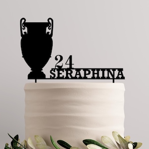 Pode incluir: Topper de bolo em silhueta preta com um troféu, o número 24 e o nome Seraphina. O topper está em um bolo branco.