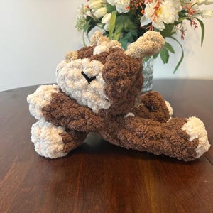 May include: A handmade, plush toy in the shape of a dog. The dog is primarily brown with cream-colored accents on its face, paws, and ears. The eyes are black. The toy is made of a soft, textured yarn.