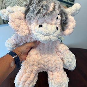 May include: A handmade Highland cow soft toy in shades of pink and white. The cow has a fluffy grey and brown faux fur mane and black button eyes. The toy is made from a textured, looped yarn, and is a cuddly design.